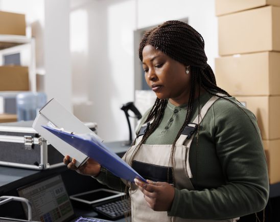 Small business worker analyzing merchandise logistics, checking customers orders on computer, preparing packages for delivery. Stockroom employee wearing industrial overall in warehouse